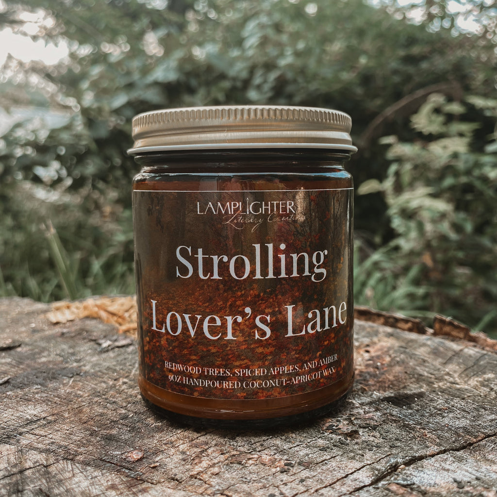 A jar candle labeled 'Strolling Lover's Lane' by Lamplighter Candle Co sits on a weathered tree stump with a blurred forest background.