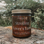 A jar candle labeled 'Strolling Lover's Lane' by Lamplighter Candle Co sits on a weathered tree stump with a blurred forest background.