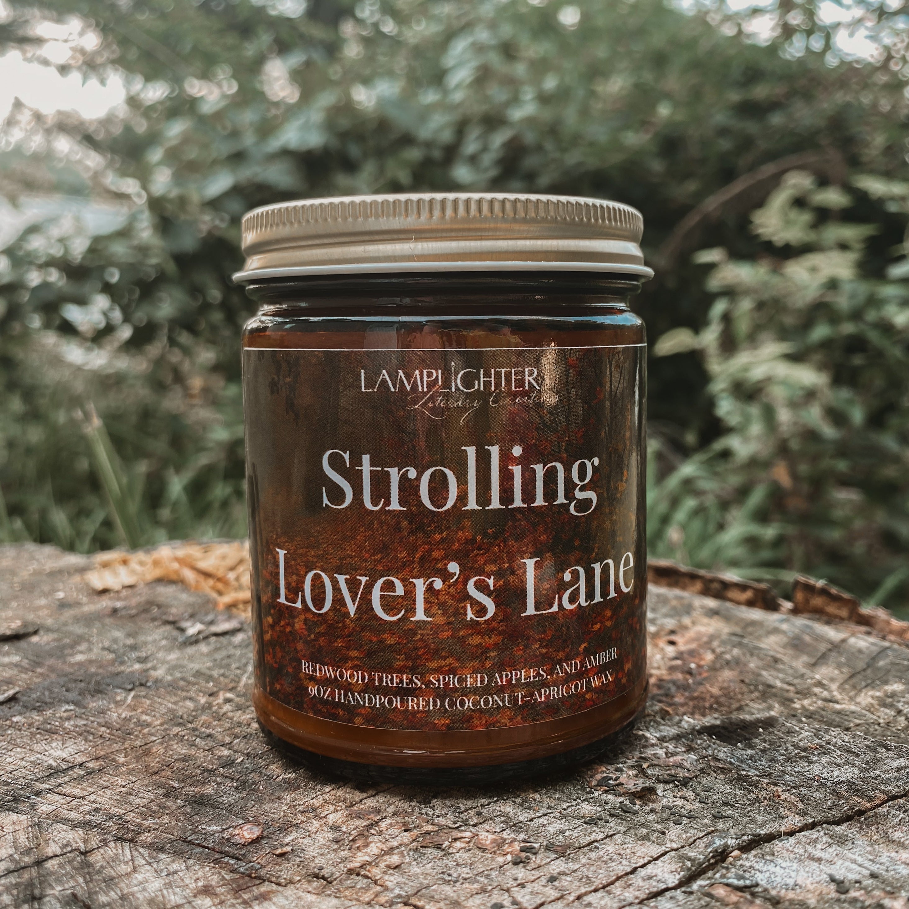 A jar candle labeled 'Strolling Lover's Lane' by Lamplighter Candle Co sits on a weathered tree stump with a blurred forest background.