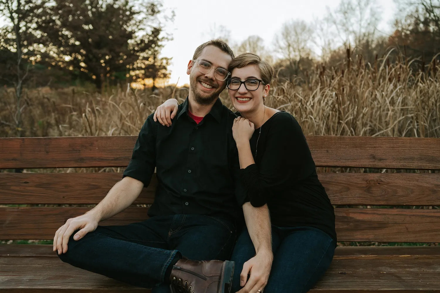 Lamplighter Literary owners Megan and Ryan sitting on a wooden bench in a park with trees and grass in the background.